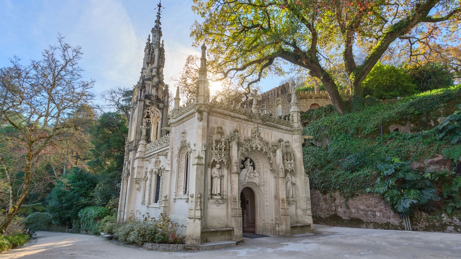 The Gothic-style chapel at Quinta da Regaleira with intricate carvings, statues, and sunlight shining through the trees.