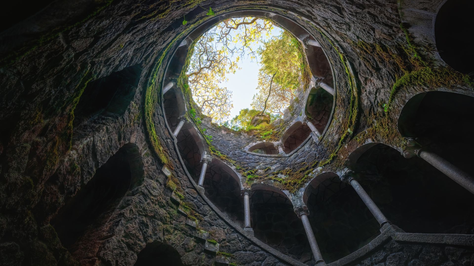 View from the bottom of the spiral Initiation Well at Quinta da Regaleira, with moss-covered stone arches and a circular opening to the sky.