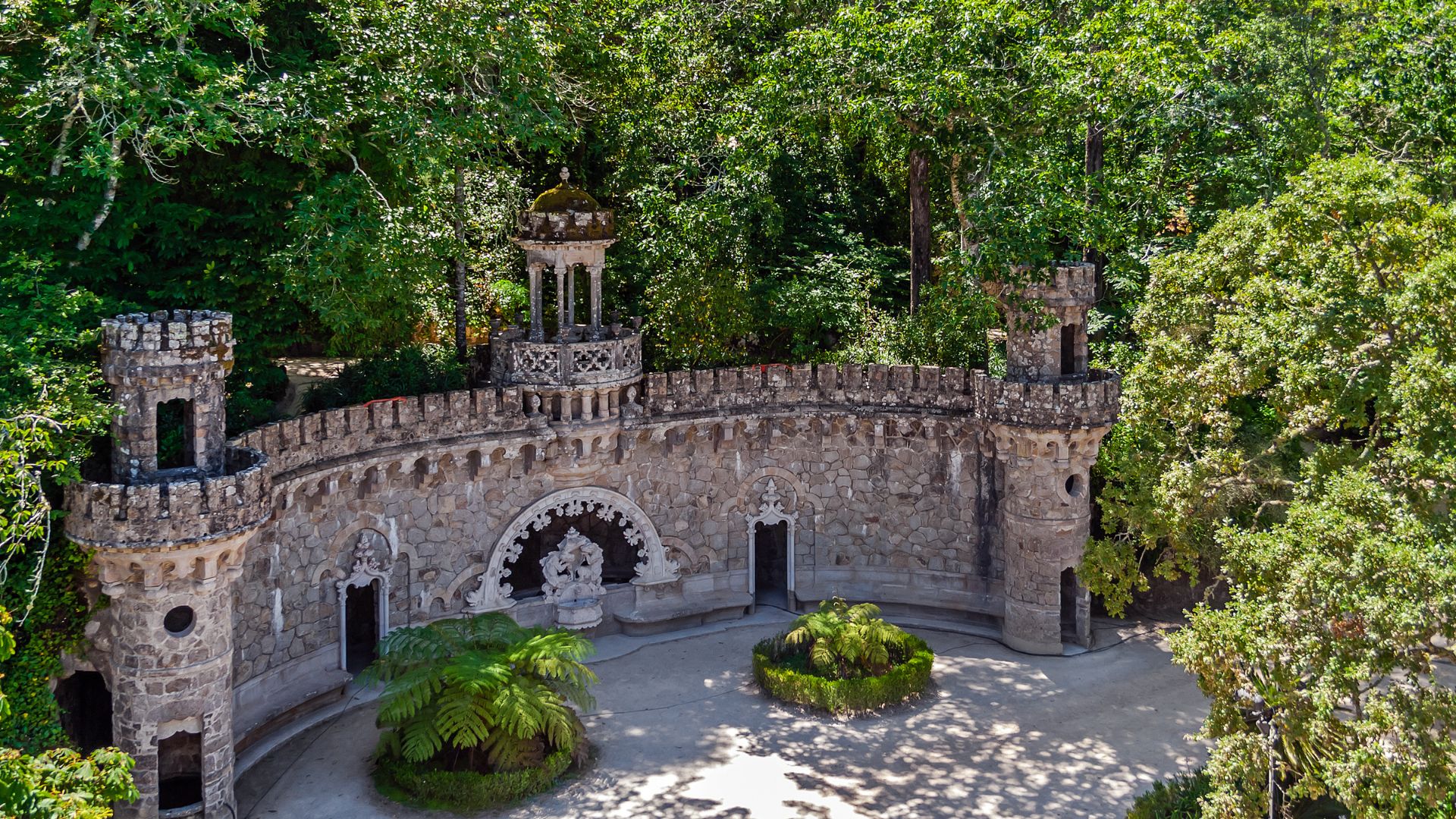 A round stone tower with decorative battlements and arches nestled in the forest at Quinta da Regaleira, surrounded by trees.