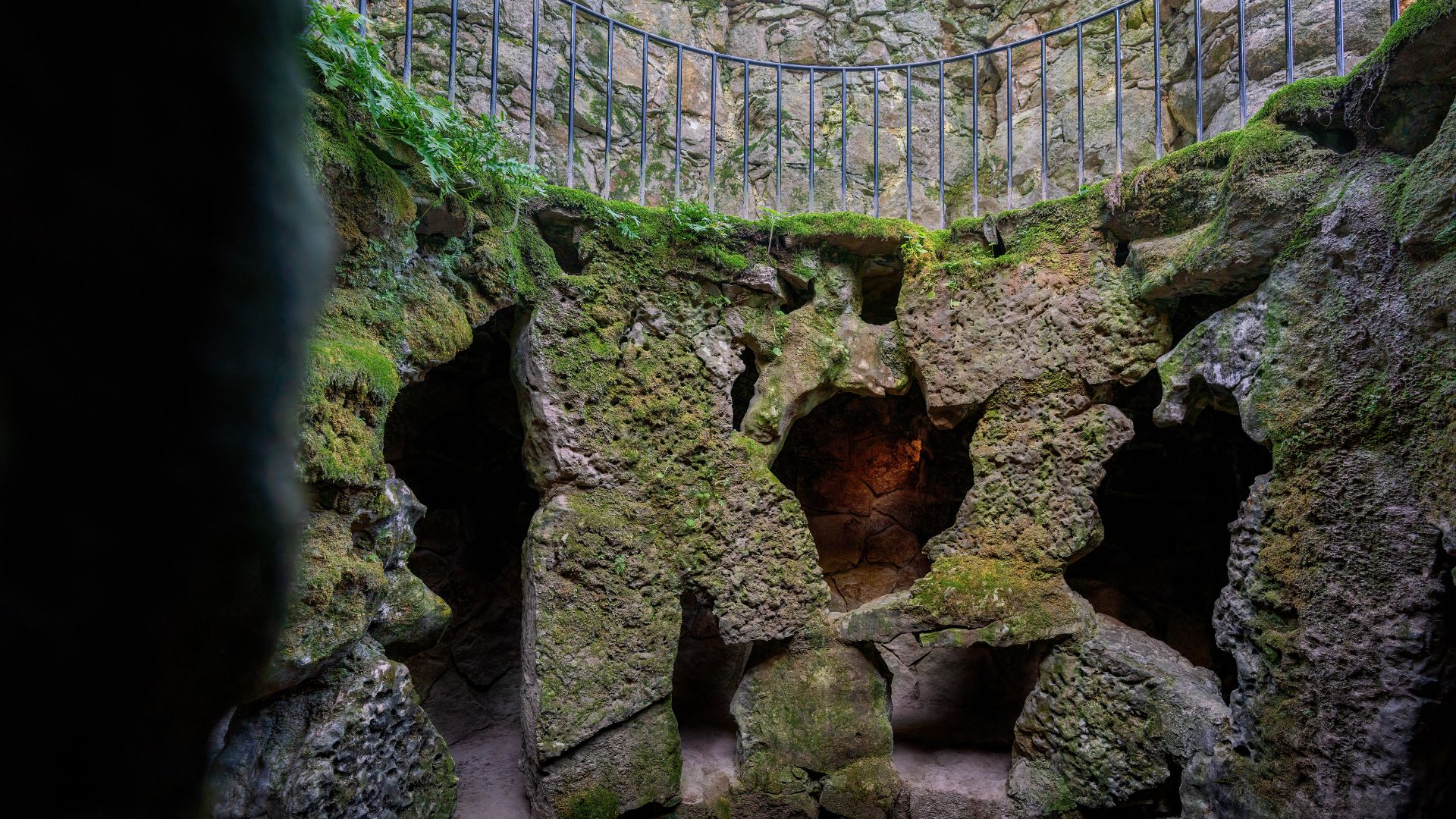 Moss-covered rock walls and passage openings inside the Unfinished Well at Quinta da Regaleira, viewed from the bottom.