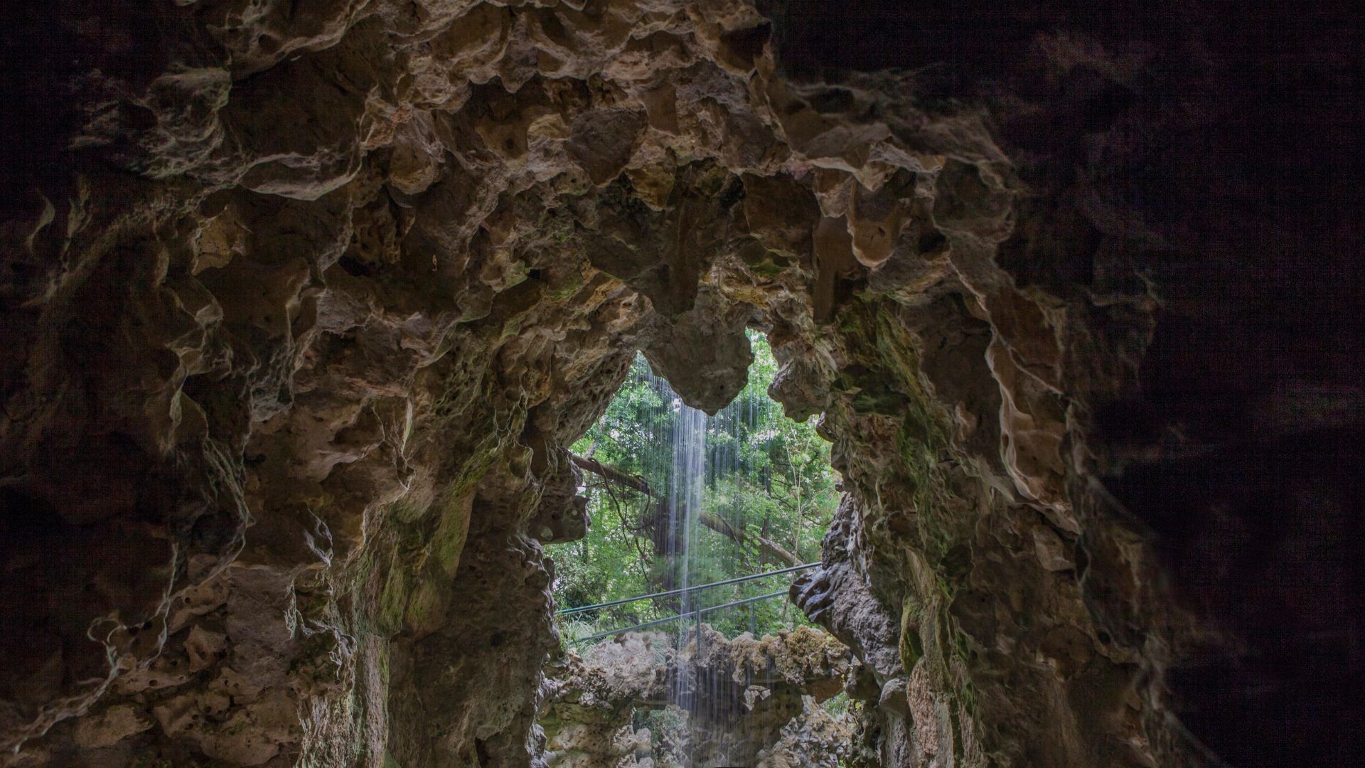 A small waterfall cascading into a lake inside a rocky cave at Quinta da Regaleira, with sunlight filtering through the foliage.